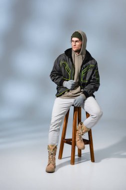 Şapkalı, anorak giyimli, gri taburede oturan modaya uygun genç bir adam. 