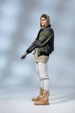 full length of young trendy man in hat, hoodie and anorak posing on grey clipart