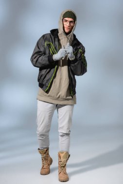 full length of young stylish man in hat, hoodie and anorak standing on grey clipart