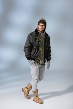 full length of trendy man in hat, hoodie and anorak posing on grey clipart