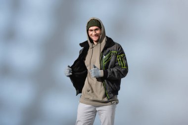 fashionable man in glasses, hat, anorak and gloves smiling and looking at camera on grey clipart