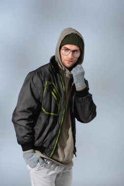 trendy man in glasses, hat, anorak and gloves standing with hand in pocket on grey clipart