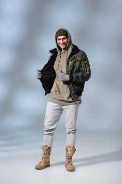full length of young cheerful man in hat, hoodie and anorak posing and showing thumb up on grey clipart