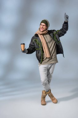 full length of excited man in hat, hoodie and anorak holding paper cup on grey clipart