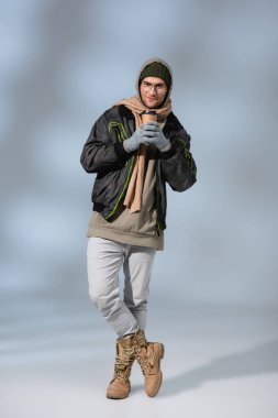 Uzun boylu, şapkalı, kapüşonlu ve anorak, bacak bacak üstüne atmış ve elinde gri bardakla bekleyen heyecanlı bir adam.