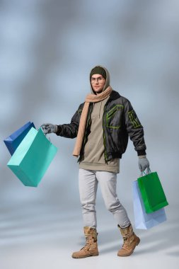 full length of stylish man in winter outfit holding shopping bags on grey clipart