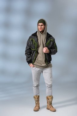 full length of stylish man in hat, hoodie and anorak standing with hand in pocket on grey clipart