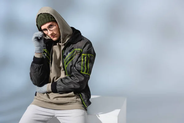 Young Trendy Man Hat Gloves Anorak Sitting White Cube While — Stock Photo, Image