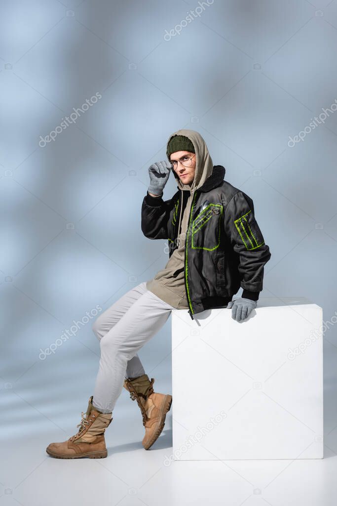 full length of trendy man in hat, gloves and parka adjusting glasses while sitting on white cube on grey 