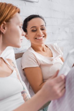 Neşeli Afrikalı Amerikalı kadın lezbiyen kız arkadaşının yanında gülümsüyor. Yatakta dijital tablet kullanıyor, ön plan bulanık.
