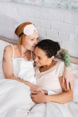 redhead lesbian woman in sleep mask on forehead embracing african american girlfriend in bed clipart