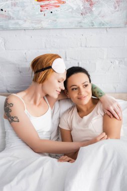 tattooed lesbian woman in eyemask on forehead hugging african american girlfriend in bed clipart