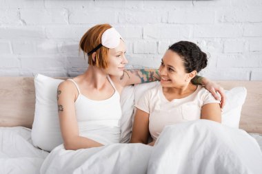 happy interracial lesbian couple looking at each other while sitting in bed clipart