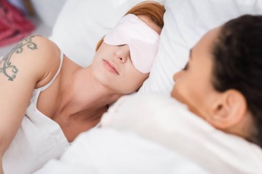 tattooed lesbian woman in eyemask sleeping near african american girlfriend on blurred foreground clipart