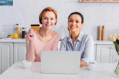 Kredi kartıyla mutlu kızıl saçlı kadın ve onun neşeli Afrikalı Amerikalı kız arkadaşı dizüstü bilgisayarın yanındaki kameraya bakıyor.