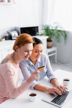 Afro-Amerikan kız arkadaşının yanında elinde kredi kartı olan şık lezbiyen bir kadın mutfaktaki dizüstü bilgisayarda yazı yazıyor.