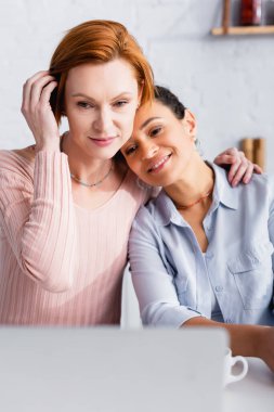 Mutfakta dizüstü bilgisayarın yanında lezbiyen Afrikalı Amerikalı kız arkadaşının omzuna sarılan kızıl saçlı kadın.