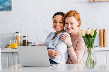 Mutfakta dizüstü bilgisayarın yanında lezbiyen Afrikalı Amerikalı kız arkadaşının omzuna sarılan neşeli kızıl kadın.