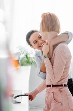 Neşeli Afro-Amerikalı kadın, bulanık ön planda, mutfakta kızıl saçlı kız arkadaşını kucaklıyor.