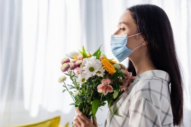 Evde elinde çiçekle gezen tıbbi maskeli genç alerjik kadının yan görüntüsü.