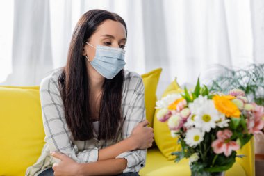 Üzgün, alerjik, maskeli bir kadın. Çiçeklerin yanında oturuyor.