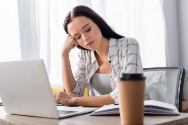exhausted freelancer sitting near laptop and coffee to go on blurred foreground clipart