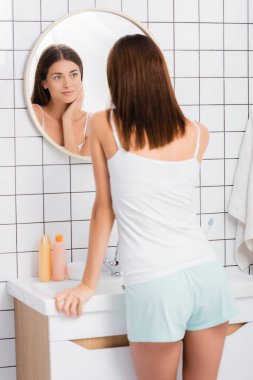 Beyaz cüppeli gülümseyen kadın banyoda dikilip aynaya bakıyor.