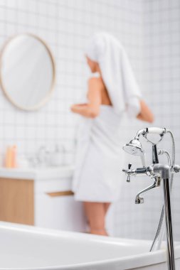 Duşlu musluğun seçici odağı ve banyoda bulanık arka planda duran beyaz havlulara sarılı kadın.