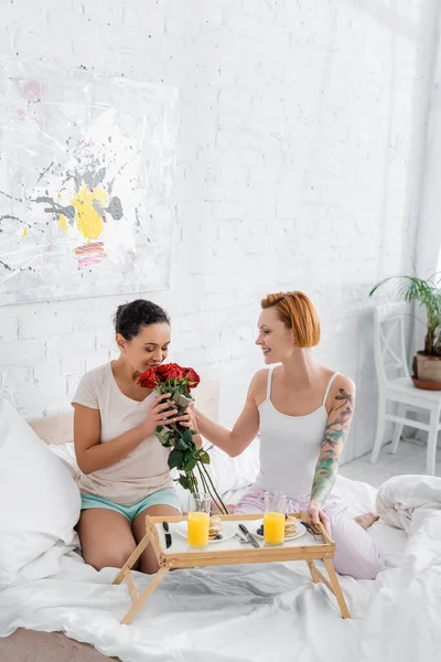 非洲裔美国女人在红头发女同志女友身边闻到红玫瑰的香味 在床上吃早餐 — 图库照片