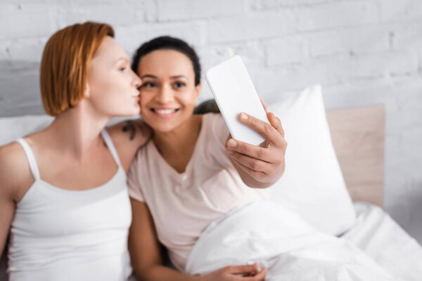 cheerful african american woman taking selfie with redhead lesbian girlfriend kissing her in bed, blurred foreground