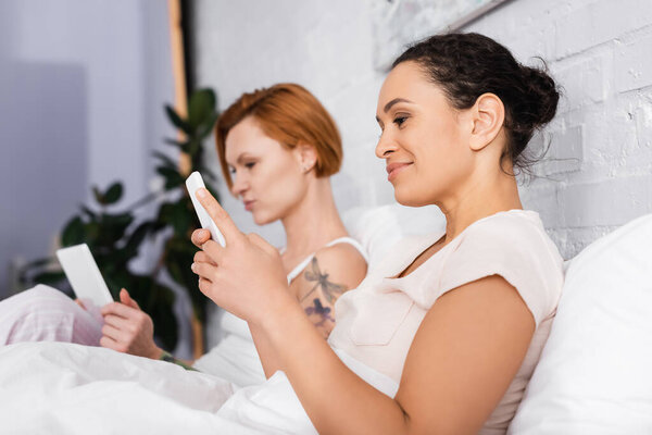 smiling african american woman using smartphone near lesbian girlfriend with laptop on blurred background