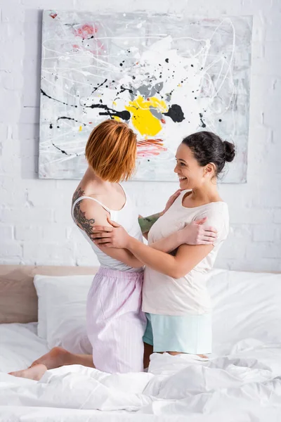 赤毛のレズビアン女性と陽気なアフリカ系アメリカ人の彼女の側面図は ベッドルームで抱き合ってお互いを見て — ストック写真