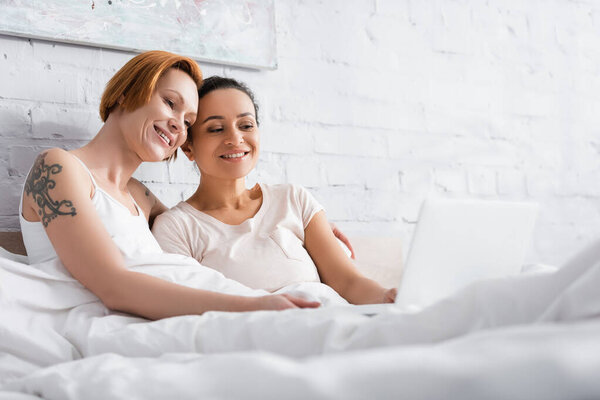 happy interracial lesbian couple watching movie on laptop in bed on blurred foreground