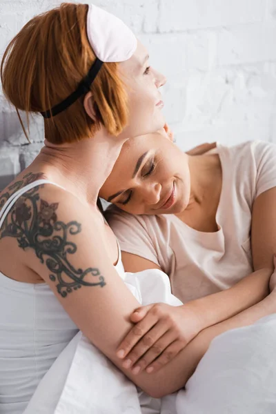 tattooed lesbian woman in sleep mask on forehead embracing african american girlfriend in bed