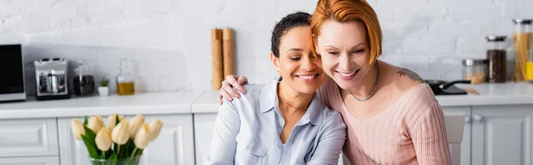 红头发女同性恋者抱着快乐的非洲女友在厨房靠近郁金香的地方挂着模糊的前景 — 图库照片