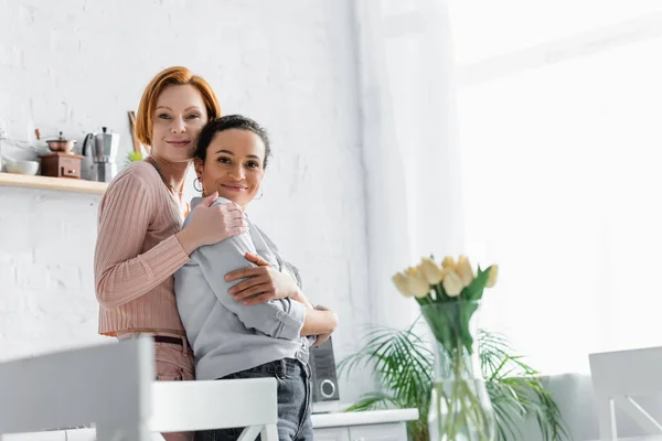 Redhead Lesbian Woman Embracing African American Girlfriend Kitchen Tulips Blurred — Stock Photo, Image