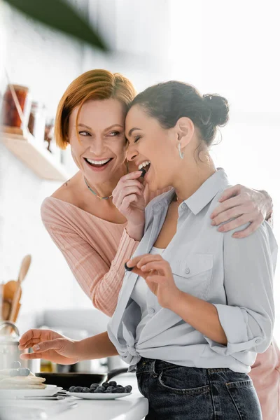 Cheerful Lesbian Woman Feeding African American Girlfriend Blueberry Kitchen — Stock Photo, Image