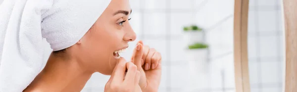 side view of woman with white towel on head flossing teeth near mirror in bathroom, banner