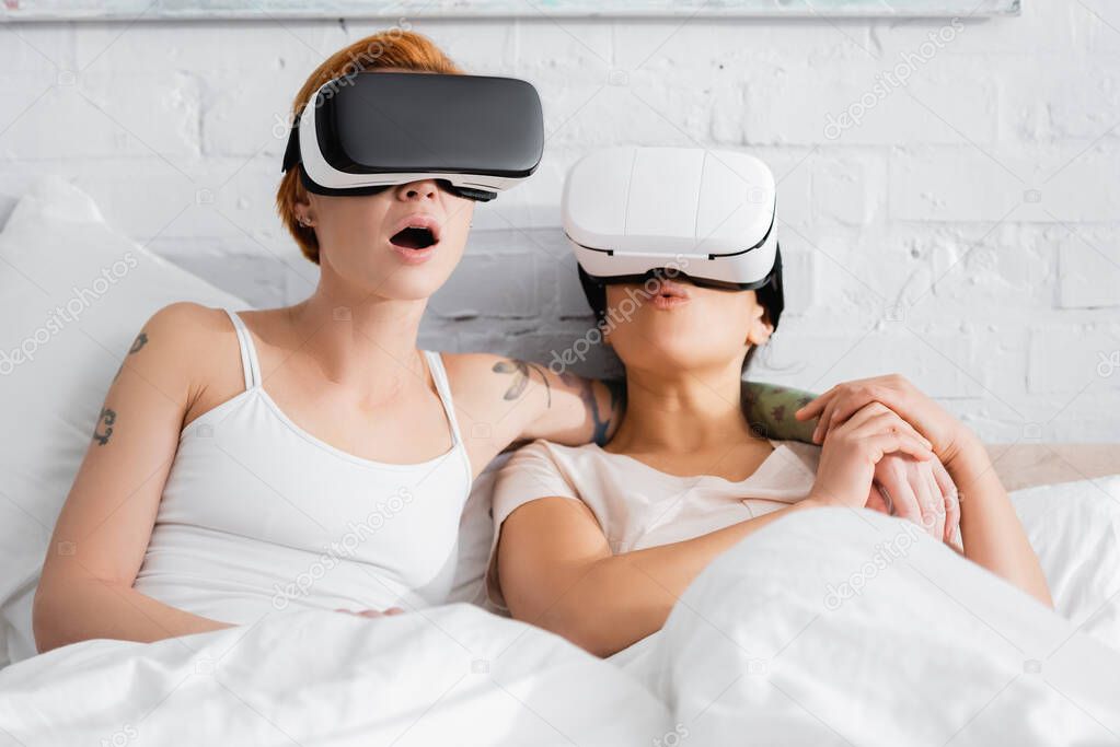 astonished lesbian woman in vr headset embracing shoulder of african american girlfriend in bed