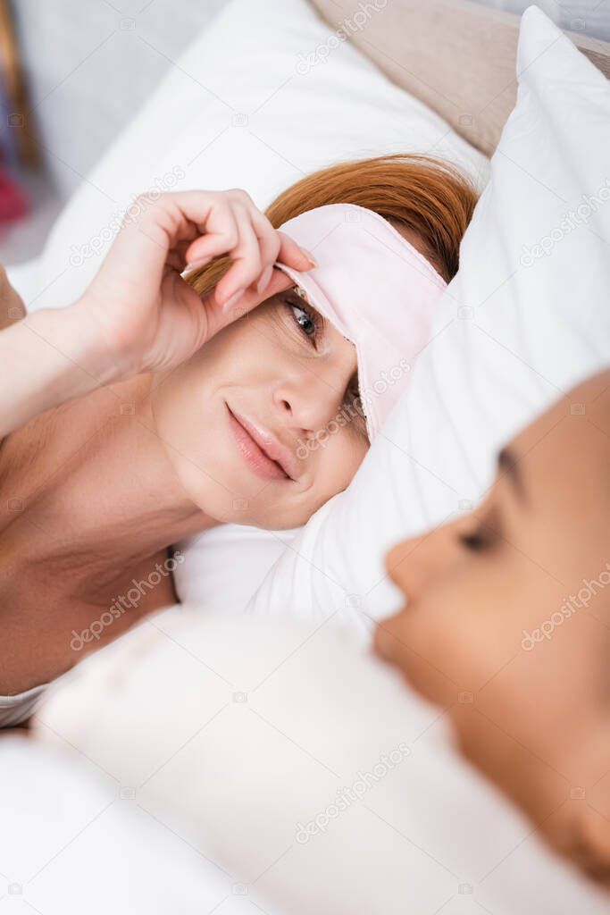 smiling lesbian woman in eyemask looking at african american girlfriend sleeping beside in bed on blurred foreground