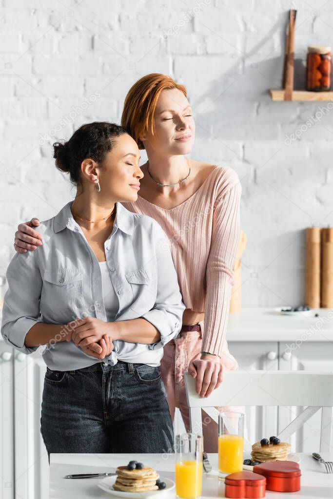 stylish interracial lesbian couple standing with closed eyes near table with breakfast and valentines day presents