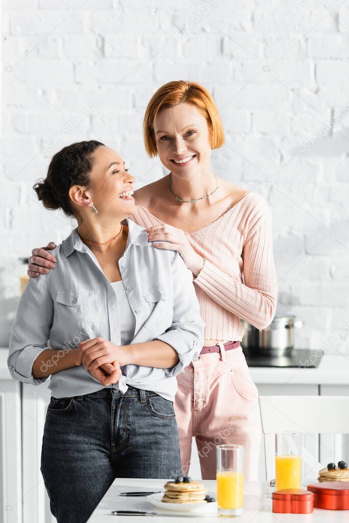 smiling lesbian woman embracing shoulders of laughing african american girlfriend near breakfast and valentines day gifts on table