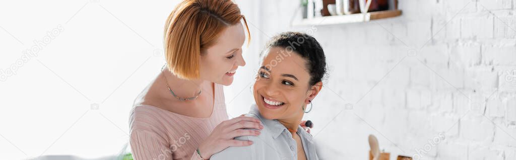cheerful interracial lesbian couple smiling while looking at each other at home, banner