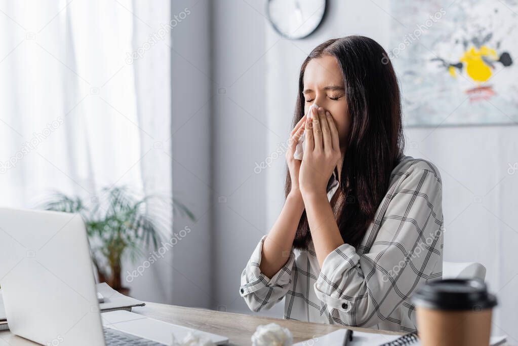 young allergic freelancer sneezing in paper napkin near laptop and coffee to go on blurred foreground