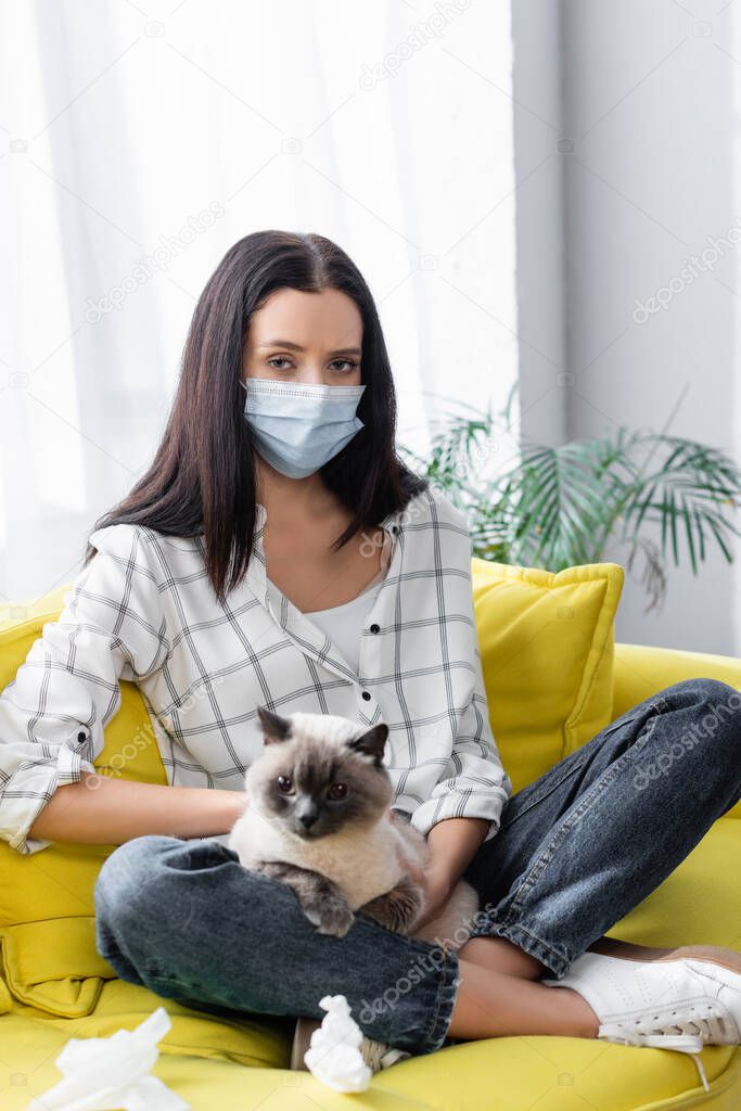 allergic woman in medical mask looking at camera while sitting on sofa with cat