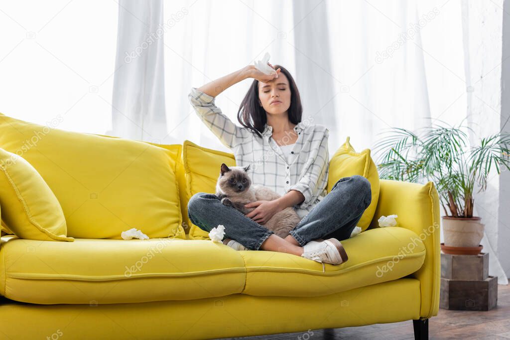 young allergic woman sitting on sofa with cat and suffering from headache
