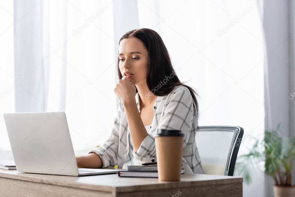 thoughtful freelancer working at laptop near coffee to go on blurred foreground