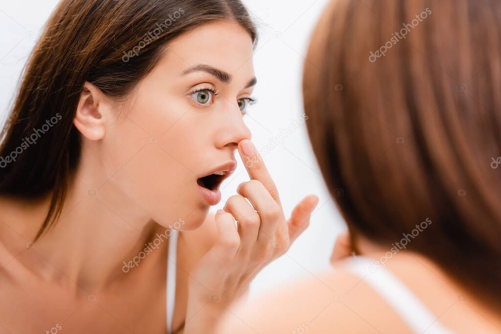 shocked young woman touching nose while looking in mirror in bathroom, blurred foreground