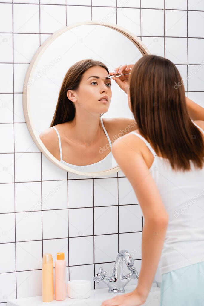 young woman in singlet tweezing eyebrows near mirror in bathroom