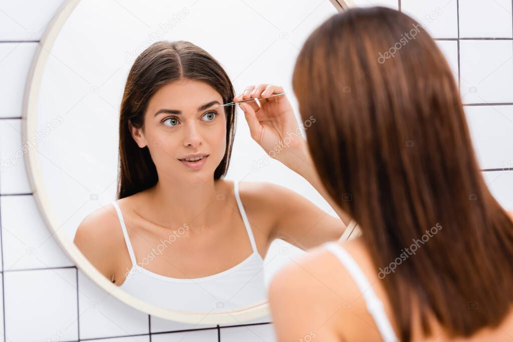 young woman in white singlet tweezing eyebrows near mirror in bathroom
